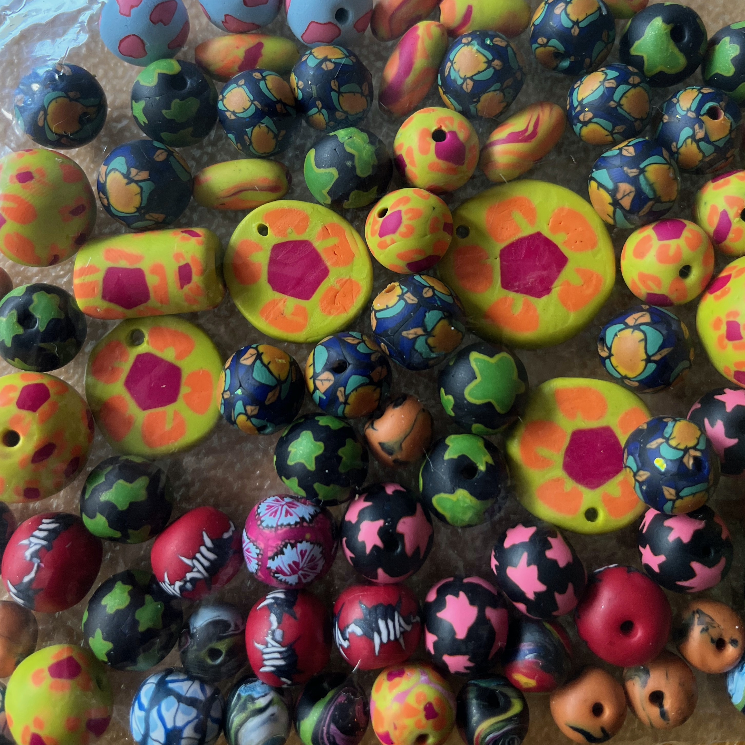 A collection of polymer clay beads in a plastic bag. The beads are all multicolored with various designs: flowers, barbed wire, and abstract marble. They are slightly obscured by the crinkly bag that contains them.