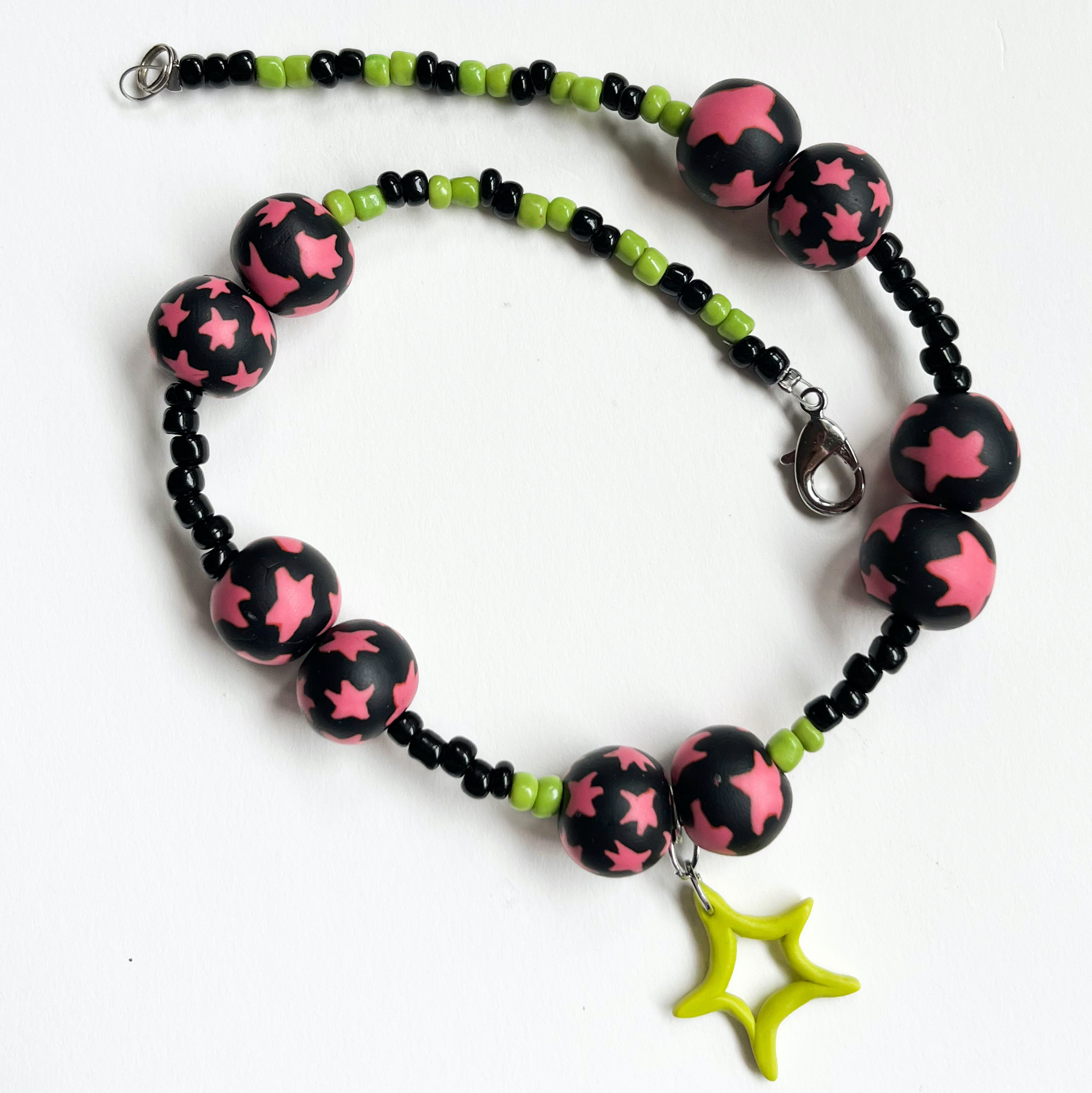 A necklace featuring handmade polymer clay beads with a pink and black color scheme, strung alongside green and black seed beads and a star-shaped pendant made from polymer clay.